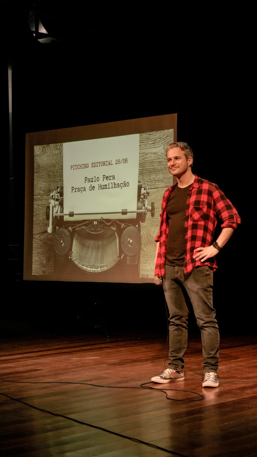 Paulo Pera — homem de pele clara, cabelos curtos, escuros e com vários fios grisalhos, vestindo camisa xadrez vermelha e preta sobre camiseta preta e calça jeans também preto — está de pé no palco durante uma apresentação, sorrindo, segurando um microfone, em um ambiente escuro. Ao fundo, um slide projetado mostra uma imagem de máquina de escrever com uma folha escrita: “Pitching Editorial 28/06. Paulo Pera. Praça de Humilhação”.