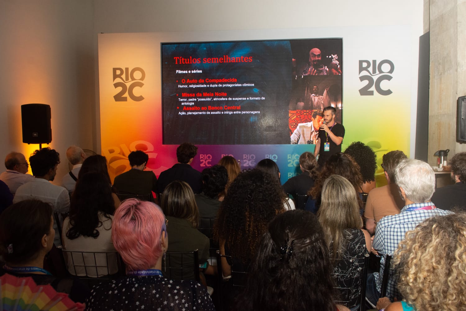 Apresentação do Pitching Editorial no evento Rio2C com uma plateia atenta assistindo o Paulo Pera — homem de pele clara, cabelos curtos e escuros, barba curta e aparada, usando uma camiseta preta. Ele está em pé, segurando um microfone com a mão direita e usando um crachá colorido do evento pendurado no pescoço — apresentando o seu projeto 'Possessão', diante de uma tela grande. Na tela, há uma apresentação com o título 